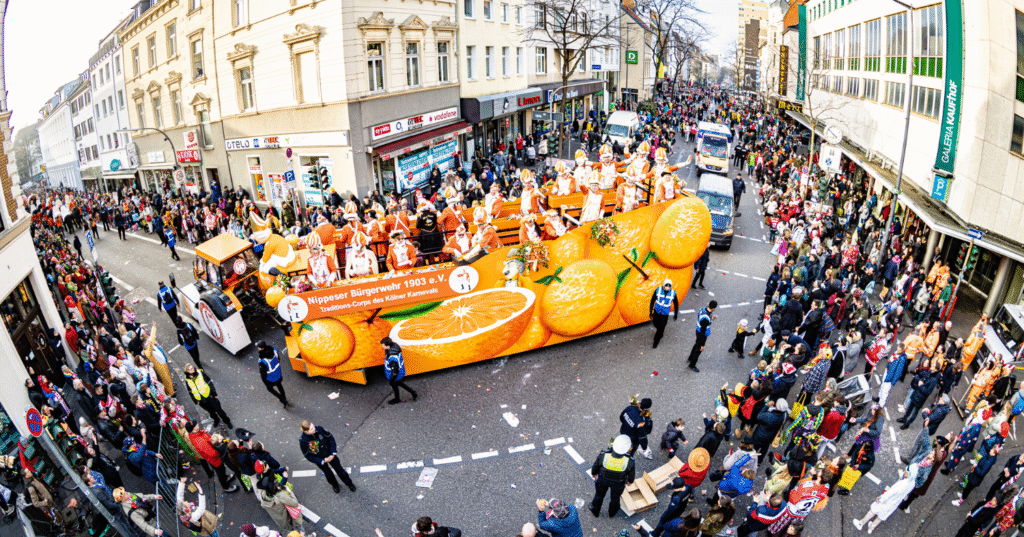 Von einer weiteren Perspektive der fahrende Apfelsinen Zugwagen der Nippeser Bürgerwehr. Drumherum stehen kostümierte Menschen.